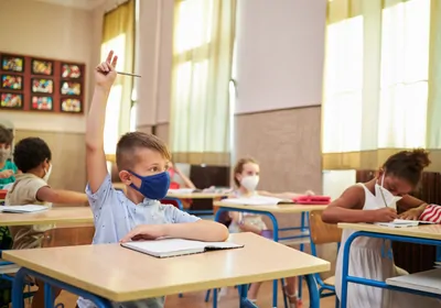masked boy sitting in classroom raising his hand